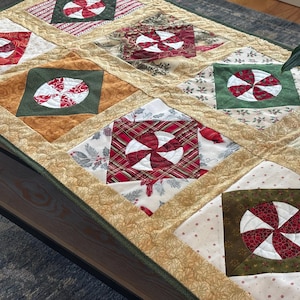 May include: A rectangular quilted table runner featuring a repeating pattern of square blocks. Each block contains a circular red and white peppermint candy design set against various fabric backgrounds including green, gold, and holiday prints.
