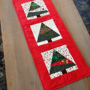 May include: A red Christmas table runner with three appliqued Christmas trees. Each tree is a different pattern of green fabric, with a brown trunk, set on a white square. The runner has a red polka dot border.