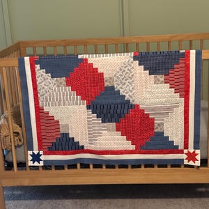 May include: A handmade quilt with a patriotic theme, draped over a wooden crib. The quilt features a patchwork design with red, white, and blue fabrics. The design includes a central diamond shape and star accents.