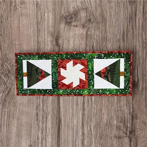 May include: A rectangular quilted table runner featuring a green border with white star accents. The center displays two triangular tree motifs in dark green and red tones on white backgrounds, separated by a central red square with a white pinwheel.