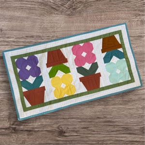 May include: A white quilted table runner with a teal border features five colorful flower pots with flowers in shades of purple, pink, yellow, and blue.