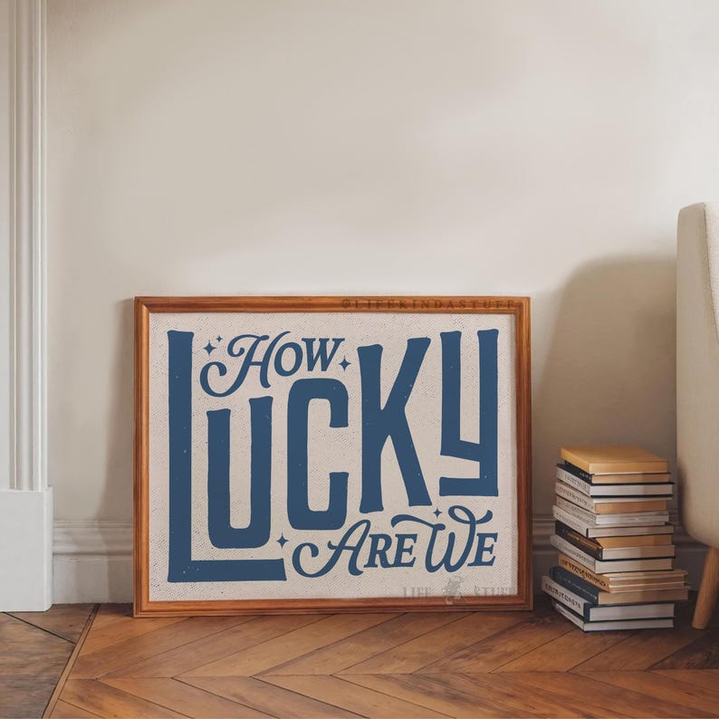 May include: A framed print with the text "How Lucky Are We" in navy blue against a beige background. The wooden frame sits on a wooden floor next to a stack of books. The print has a vintage aesthetic.