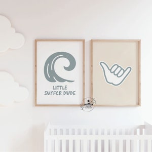 May include: Two framed prints with a surfing theme. One print features a stylized wave with the text "LITTLE SURFER DUDE" in a light blue color. The other print displays a light blue shaka hand gesture on a beige background.
