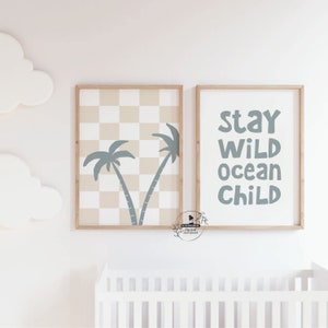 May include: Two framed wall art prints. One features a checkered pattern with palm tree silhouettes. The other print has the words "Stay Wild Ocean Child" in blue. Both are in wooden frames, suitable for a nursery or child's room.