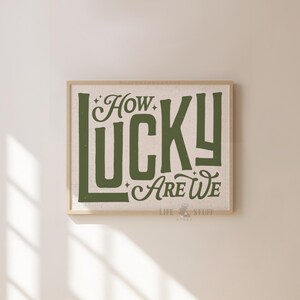 May include: A framed print with the words "How Lucky Are We" in a green, vintage-style font. The print has a beige background and a light brown wooden frame. The words are stylized with decorative elements.
