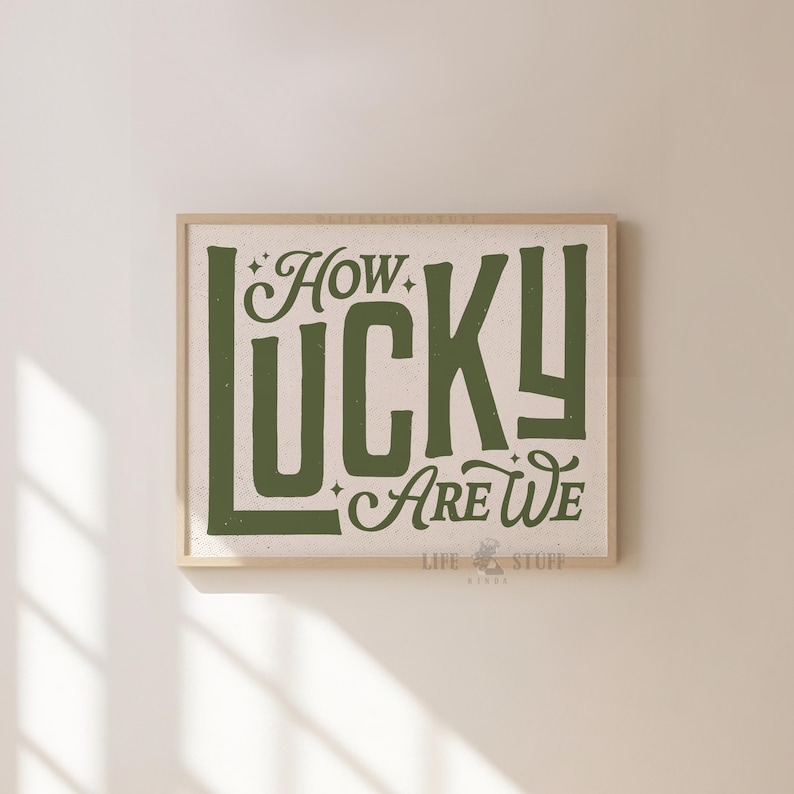 May include: A framed print with the words "How Lucky Are We" in a green, vintage-style font. The print has a beige background and a light brown wooden frame. The words are stylized with decorative elements.