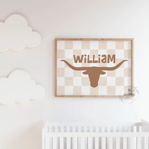 May include: A printable nursery wall art design featuring a brown longhorn steer silhouette with the name "William" printed above it. The background is a beige and white checkered pattern.