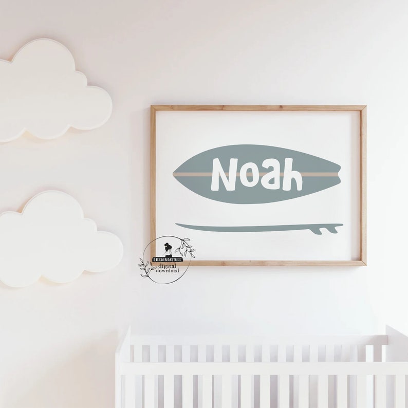 May include: A framed print featuring a teal surfboard design with the name "Noah" in white. Below the surfboard is a matching teal surfboard outline. The print is set against a white background, with decorative cloud cutouts and a crib visible.