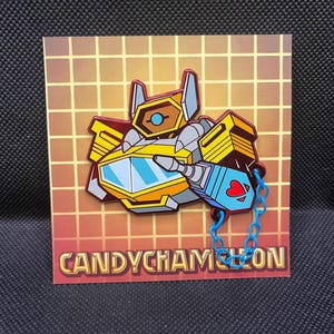 May include: A colorful pin featuring a robot design with yellow, gray, and blue accents. The pin has a blue chain and a heart detail. The pin is on a card with the text "CANDYCHAMELEON".