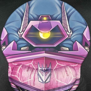 Pode incluir: Tapete de rato roxo e rosa com uma ilustração de personagem robô. O design inclui um grande elemento central em forma de olho com um brilho amarelo e um símbolo Decepticon. O tapete de rato tem um apoio para o pulso.