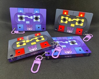 May include: Four rectangular acrylic keychains shaped like cassette tapes. Two are purple with blue accents, and two are grey with red and yellow accents. Each features a purple Decepticon logo and a light purple lobster claw clasp attached.