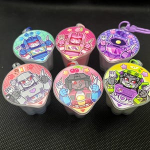 May include: Six colorful, plastic container keychains with cartoon robot designs. Each container has a different robot character on the lid, with a matching color scheme. The keychains have a small loop for attaching to keys or bags.