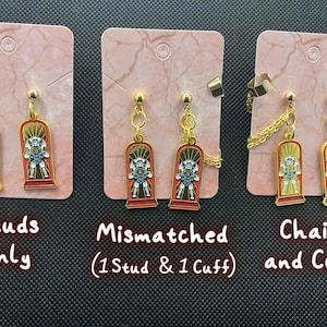 May include: Gold-toned earrings with a red and yellow design featuring a figure in a rectangular frame. The earrings are displayed on pink cards, with options for studs, mismatched, and chains with a cuff.