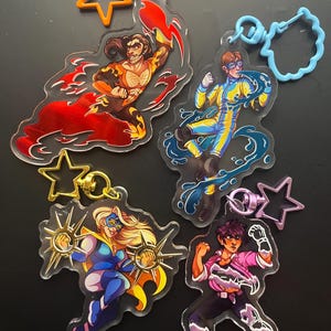 Puede incluir: Cuatro llaveros acrílicos con ilustraciones de superhéroes. Cada llavero presenta un diseño de personaje diferente, con colores vibrantes y poses dinámicas. Los llaveros tienen cierres en forma de estrella o únicos en naranja, dorado y morado.