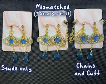 May include: Gold-coloured mismatched earrings with blue detailing. The earrings have a geometric design with a central blue gem and dangling blue teardrop gems. One pair is studs only, the other has chains and a cuff.