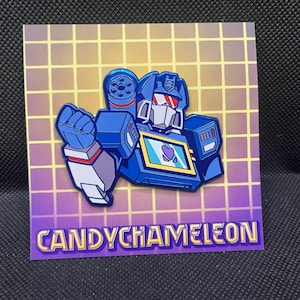 Puede incluir: Pin de robot azul con cara blanca y visera roja, sobre un fondo de cuadrícula morada y amarilla. El pin presenta un diseño en forma de corazón en el pecho y el texto "CANDYCHAMELEON" en la parte inferior.