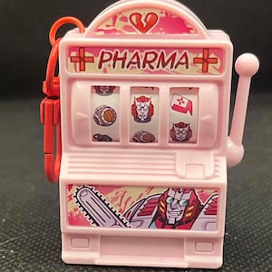 May include: A pink slot machine keychain with the word "PHARMA" and a broken heart graphic. The reels display cartoon images. A red clip is attached to the top. The bottom of the machine features a cartoon character with a chainsaw.