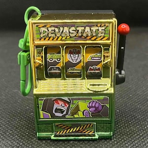 May include: A green and gold slot machine keychain with the word "DEVASTATE" across the top. The slot machine has three reels with vehicle and robot graphics. A red ball sits atop the pull lever.