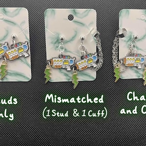 May include: Earrings with a ray gun design in white, yellow, and blue, accented with green lightning bolt charms. Available as studs, mismatched (stud and cuff), or chains and cuff styles. The earrings are displayed on white cards with a green marble pattern.