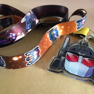 Puede incluir: Un cordón con un patrón repetido de naves espaciales de dibujos animados en tonos morados, azules y rojos sobre un fondo marrón. Un llavero acrílico transparente con un diseño de cara de robot está unido al cordón.