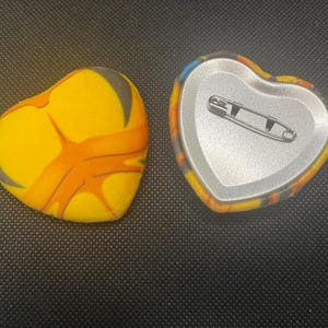 Puede incluir: Dos chapas en forma de corazón con un vibrante patrón jaspeado en amarillo y naranja. Una chapa muestra el diseño frontal, mientras que la otra está dada la vuelta para mostrar la parte trasera de metal plateado con un cierre de imperdible.