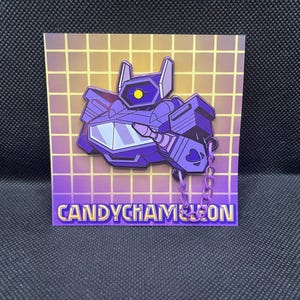May include: A purple robot pin with a yellow eye and a chain, set against a grid background. The pin features the text "CANDYCHAMELEON" in gold letters. The robot design is stylized and geometric.