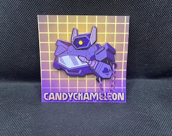 Könnte beinhalten: Ein lila Roboter-Anstecknadel mit gelbem Auge und Kette, vor einem Gitterhintergrund. Die Anstecknadel trägt den Text "CANDYCHAMELEON" in goldenen Buchstaben. Das Robot-Design ist stilisiert und geometrisch.