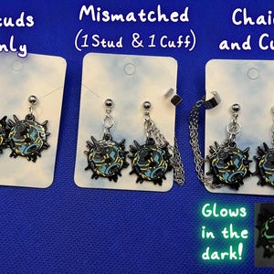 Puede incluir: Pendientes con un diseño negro, azul y amarillo, disponibles como tachuelas, desparejados (un pendiente y un puño), o con cadenas y un puño. El texto "Glows in the dark!" también es visible. Los pendientes se muestran en tarjetas blancas sobre un fondo azul.