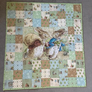 May include: A patchwork quilt featuring a central illustration of two rabbits, one carrying a sack and the other holding vegetables. The quilt is constructed from square fabric panels in shades of light green, blue, and tan, decorated with various Peter Rabbit motifs and patterns, finished with a light green border.