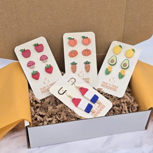 May include: A variety of colorful handmade earrings in a cardboard box. The earrings include fruit and vegetable designs like apples, peaches, carrots, and avocados. One pair of earrings is designed as red, white, and blue popsicles. The brand name "Mugs 'N Things Mercantile" is printed on the cards.