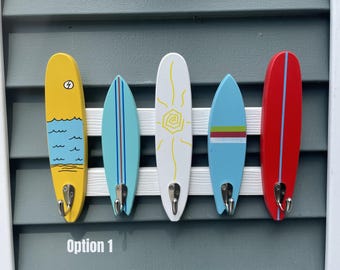 Puede incluir: Un perchero de pared con cinco placas de madera en forma de tabla de surf en amarillo, azul claro, blanco, azul medio y rojo, cada una con un gancho de metal plateado. El texto "Option 1" está impreso en blanco en la parte inferior.