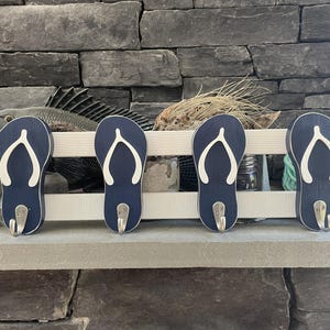 May include: Four navy blue flip-flop shaped wall hooks with white detailing and silver hooks. The hooks are mounted on a white wooden board. The background features a stone fireplace and decorative items.