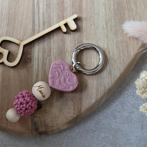 Könnte beinhalten: Ein Schlüsselanhänger mit einem herzförmigen rosa Anhänger und einem Perlenakzent. Der Herzanhänger hat ein Blättermuster. Der Schlüsselanhänger hat auch eine Holzperle mit dem Wort "Mama".