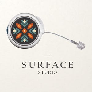 Może przedstawiać: Srebrne urządzenie Surface Studio z dekoracyjnym, czarnym, pomarańczowym i turkusowym okrągłym wzorem. Urządzenie ma cienki, srebrny kabel, który rozciąga się do przezroczystego złącza. Poniżej urządzenia znajduje się tekst "SURFACE STUDIO".