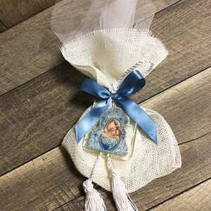 May include: A decorative favor bag made of burlap and tulle, tied with a blue satin ribbon. It features a religious icon and two white tassels. The bag is presented on a wooden surface.