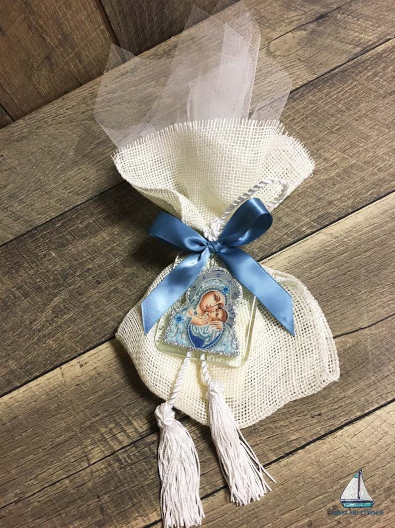 May include: A decorative favor bag made of burlap and tulle, tied with a blue satin ribbon. It features a religious icon and two white tassels. The bag is presented on a wooden surface.