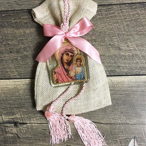 May include: A burlap bag tied with a pink satin ribbon and a decorative tassel. A clear, rectangular charm with a religious image of a woman and child is attached to the bag. The charm has gold glitter trim and pink accents.
