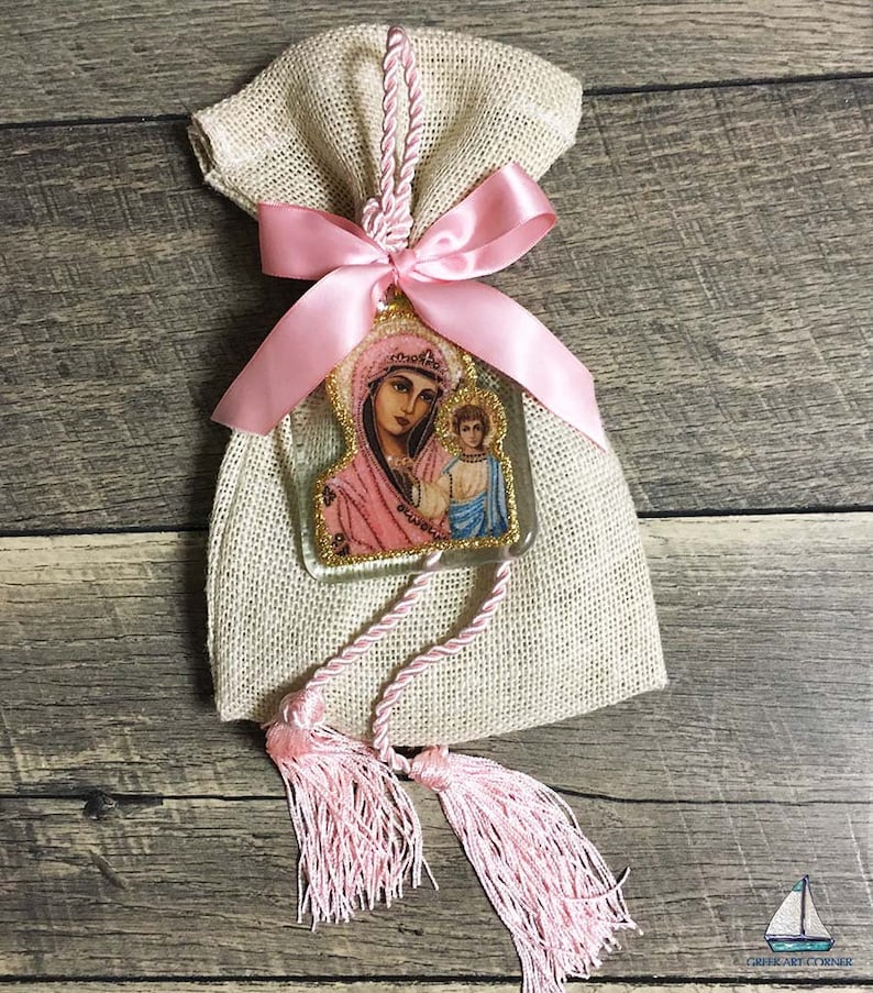 May include: A burlap bag tied with a pink satin ribbon and a decorative tassel. A clear, rectangular charm with a religious image of a woman and child is attached to the bag. The charm has gold glitter trim and pink accents.