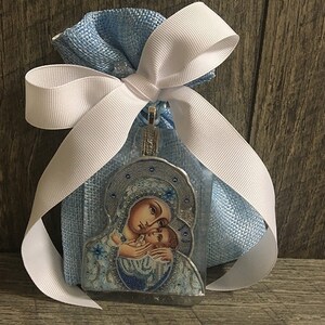 May include: A religious icon pendant featuring a mother and child, presented in a light blue mesh bag tied with a white ribbon. The icon has a silver-colored frame and is decorated with blue and white details. The background is a wooden surface.