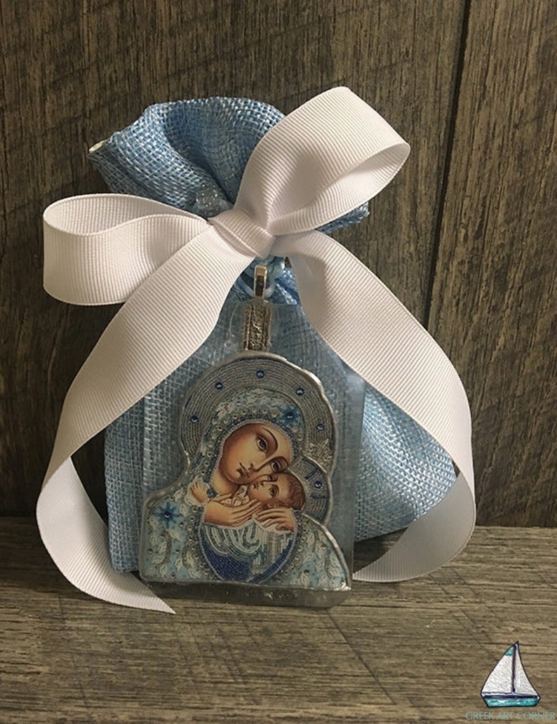 May include: A religious icon pendant featuring a mother and child, presented in a light blue mesh bag tied with a white ribbon. The icon has a silver-colored frame and is decorated with blue and white details. The background is a wooden surface.