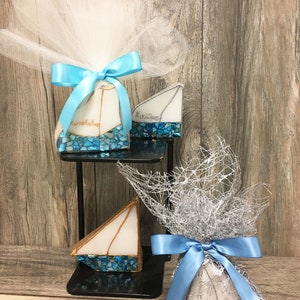 May include: Three small, blue and white mosaic sailboats with a white sail, wrapped in blue and white tulle and tied with blue ribbon. The sailboats are on a black stand.