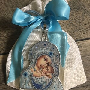 May include: A religious pendant featuring an image of the Virgin Mary and baby Jesus, presented in a white mesh bag tied with a light blue satin ribbon. The pendant has a silver-colored border and a clear, protective coating.
