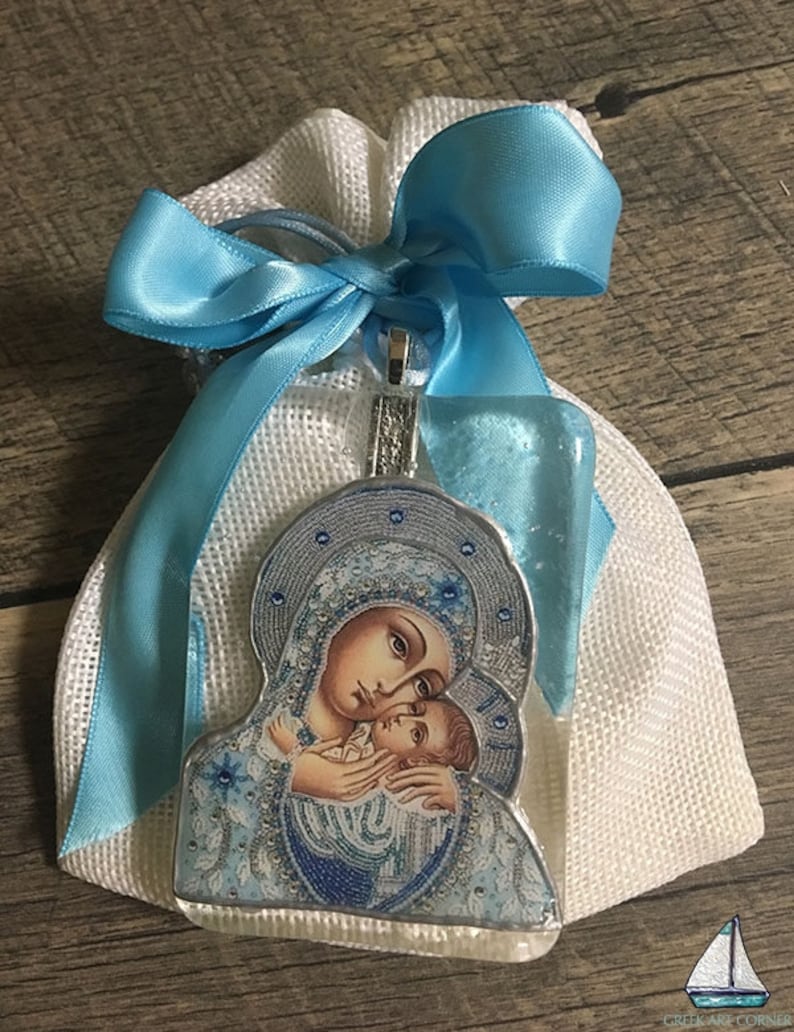 May include: A religious pendant featuring an image of the Virgin Mary and baby Jesus, presented in a white mesh bag tied with a light blue satin ribbon. The pendant has a silver-colored border and a clear, protective coating.