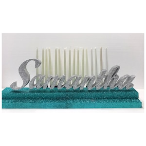 May include: Silver glitter letters spelling "Samantha" on a turquoise glitter base with white candles.