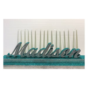 May include: Teal and silver glitter candle holder with the name "Madison" in silver glitter letters. The holder has 18 white candles.