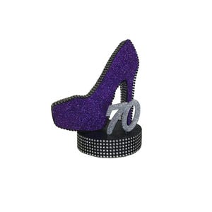 Pu&ograve; includere: Una scarpa con tacco alto glitterata viola con il numero '70' argentato, posta su una base nera. La base e il bordo della scarpa sono adornati con strass argentati. La scarpa &egrave; un oggetto decorativo.