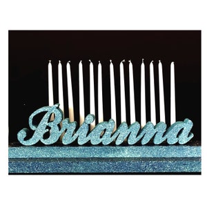 May include: A teal glittery name, "Brianna", sits in front of a row of white candles. The name is on a dark blue glittery base. The background is black.