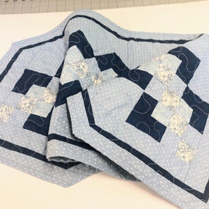 May include: A blue and white quilted table runner with a navy blue border and a floral pattern. The runner is folded in half and has a white background with blue polka dots.