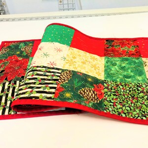 May include: A Christmas-themed quilted table runner with a red border. The runner features various patterned squares, including poinsettias, snowflakes, pinecones, and holly. The color palette includes red, green, gold, black, and white.