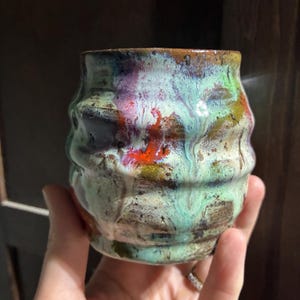 Op de afbeelding: Een keramieken beker met een golvende, geribbelde textuur en een glanzend glazuur met een gemarmerde mix van mintgroen, dieppaars, feloranje en aardse bruintinten. Het oppervlak heeft een reactieve afwerking met zichtbare druppels en kleurvariaties.